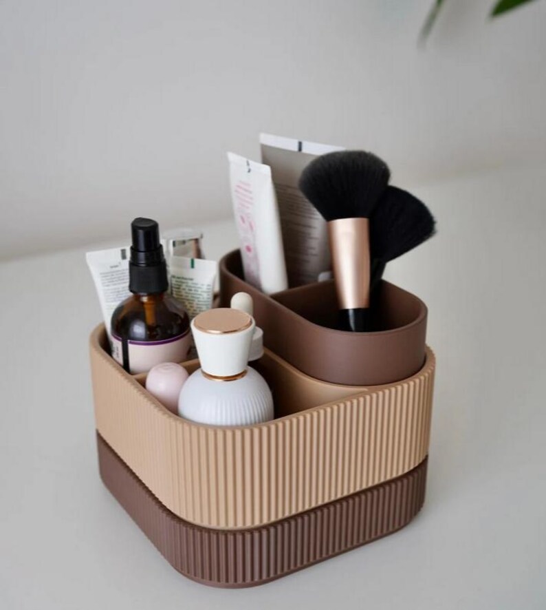 May include: A two-tiered, brown and beige cosmetic organizer with vertical ribbed detailing. The organizer holds various beauty products, including brushes, tubes, and bottles. The top tier is a lighter beige, while the bottom tier is a darker brown.