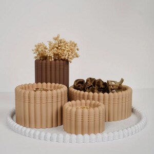 May include: A set of three beige fluted storage containers and one dark brown fluted cylindrical vase containing dried flowers, arranged on a white circular tray with a beaded edge. The containers hold small items like hair accessories.