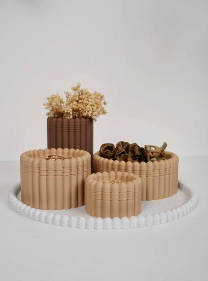 May include: A set of three beige fluted storage containers and one dark brown fluted cylindrical vase containing dried flowers, arranged on a white circular tray with a beaded edge. The containers hold small items like hair accessories.