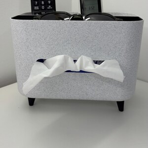 Op de afbeelding: Een gespikkelde grijze tissuebox met een rechthoekige opening waar witte tissues uitkomen. De doos heeft zwarte poten en bevat een afstandsbediening en een kleine digitale klok. Zonnebrillen liggen bovenop de doos.