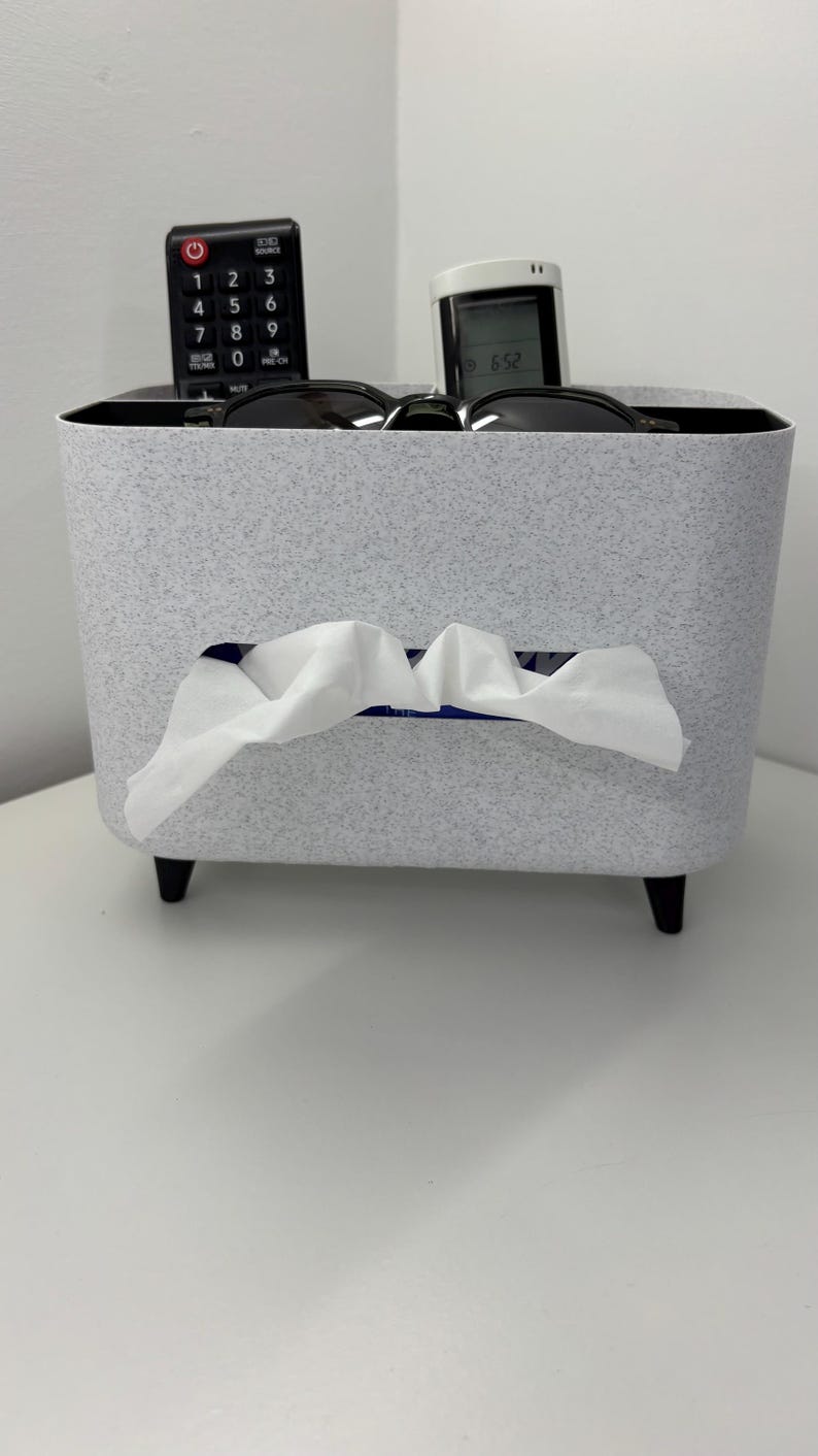 Op de afbeelding: Een gespikkelde grijze tissuebox met een rechthoekige opening waar witte tissues uitkomen. De doos heeft zwarte poten en bevat een afstandsbediening en een kleine digitale klok. Zonnebrillen liggen bovenop de doos.