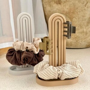 May include: Two arched, vertical stands designed for hair accessories. One stand is light gray, holding a beige ribbed scrunchie and a dark brown textured scrunchie. The other stand is tan, holding a waffle-textured scrunchie and two hair clips.