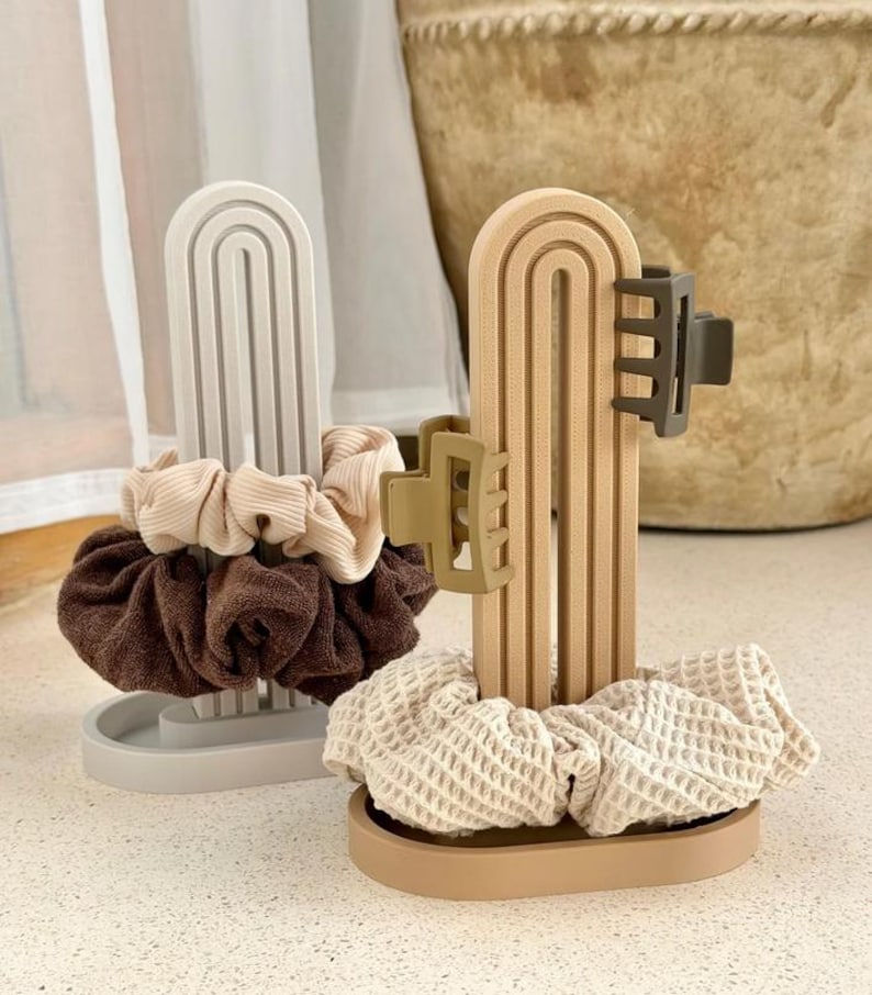 May include: Two arched, vertical stands designed for hair accessories. One stand is light gray, holding a beige ribbed scrunchie and a dark brown textured scrunchie. The other stand is tan, holding a waffle-textured scrunchie and two hair clips.