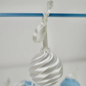 Pode incluir: Um ornamento esf&eacute;rico branco com uma textura nervurada em espiral pendurado numa fita de cetim branca. O ornamento est&aacute; suspenso numa barra horizontal azul fina, com ornamentos azuis e brancos adicionais vis&iacute;veis no fundo desfocado.