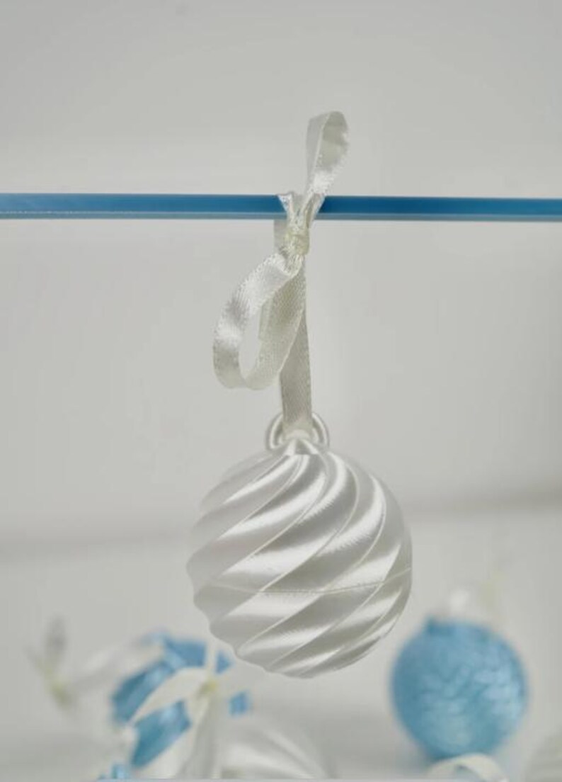Pode incluir: Um ornamento esf&eacute;rico branco com uma textura nervurada em espiral pendurado numa fita de cetim branca. O ornamento est&aacute; suspenso numa barra horizontal azul fina, com ornamentos azuis e brancos adicionais vis&iacute;veis no fundo desfocado.