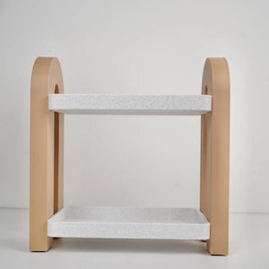 May include: A two-tiered decorative shelf with a light tan frame and speckled white shelves. The frame has an arched top and rectangular supports. The shelves are rectangular and appear to be made of a composite material.