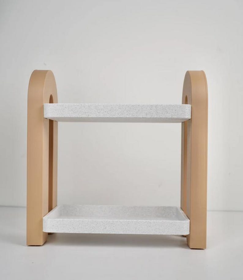 May include: A two-tiered decorative shelf with a light tan frame and speckled white shelves. The frame has an arched top and rectangular supports. The shelves are rectangular and appear to be made of a composite material.