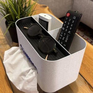 Op de afbeelding: Een gespikkelde grijszwarte organizer met meerdere compartimenten. Het bevat een afstandsbediening, zonnebril, een klein wit apparaat en een doos tissues. De organizer staat op een houten tafel.