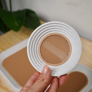 May include: A round, light gray coaster with a textured, ridged edge and a tan mesh center. The coaster is held by a hand, with a matching coaster and tray visible in the background. The coaster is made of a durable material.