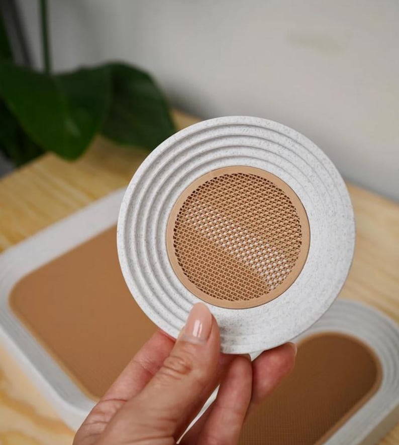 May include: A round, light gray coaster with a textured, ridged edge and a tan mesh center. The coaster is held by a hand, with a matching coaster and tray visible in the background. The coaster is made of a durable material.
