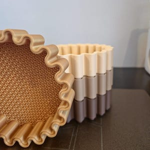 May include: A set of three-tone, wavy-edged containers in shades of beige, tan, and brown. The containers are stacked, with a textured interior visible in the open container. The design suggests a decorative or organizational purpose.