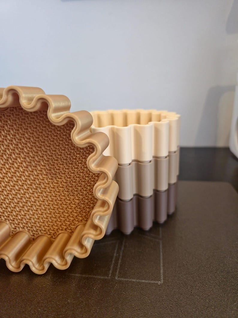 May include: A set of three-tone, wavy-edged containers in shades of beige, tan, and brown. The containers are stacked, with a textured interior visible in the open container. The design suggests a decorative or organizational purpose.