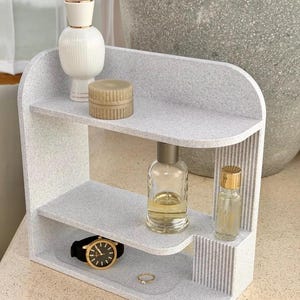 May include: A light gray speckled two-tier shelf with rounded edges and a vertical ribbed side panel. The top shelf holds a white bottle and a round tan container. The bottom shelf holds a clear glass bottle, a small vial, a watch, and a ring.