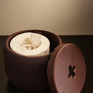 May include: A brown, ribbed toilet paper holder with a matching lid. The lid is open, revealing a roll of white toilet paper. The holder has a button-shaped design on the lid.
