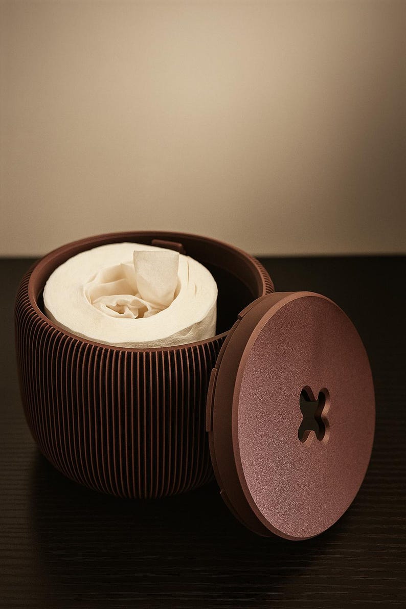 May include: A brown, ribbed toilet paper holder with a matching lid. The lid is open, revealing a roll of white toilet paper. The holder has a button-shaped design on the lid.