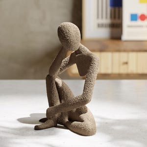 May include: A beige textured sculpture of a person in a seated, contemplative pose. The figure rests their chin on one hand, with the other hand resting on their knee. The sculpture has a rough, granular surface and is placed on a white surface.