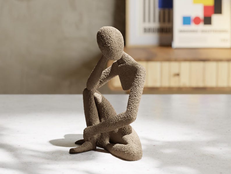 May include: A beige textured sculpture of a person in a seated, contemplative pose. The figure rests their chin on one hand, with the other hand resting on their knee. The sculpture has a rough, granular surface and is placed on a white surface.