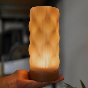 Może przedstawiać: Świecąca, teksturowana lampa o falistym wzorze. Lampa ma ciepłą, bursztynową poświatę i brązową podstawę. Pionowe linie tworzą efekt żebrowania na powierzchni lampy. Lampa jest trzymana w dłoni.