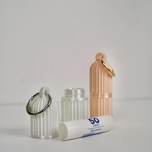 May include: Two cylindrical, ribbed, white and peach-colored plastic cases with attached metal rings, alongside a white lip balm tube labeled with the number 50 and the word HOCH, featuring a small blue icon and the text UVA + UVB SCHUTZ.