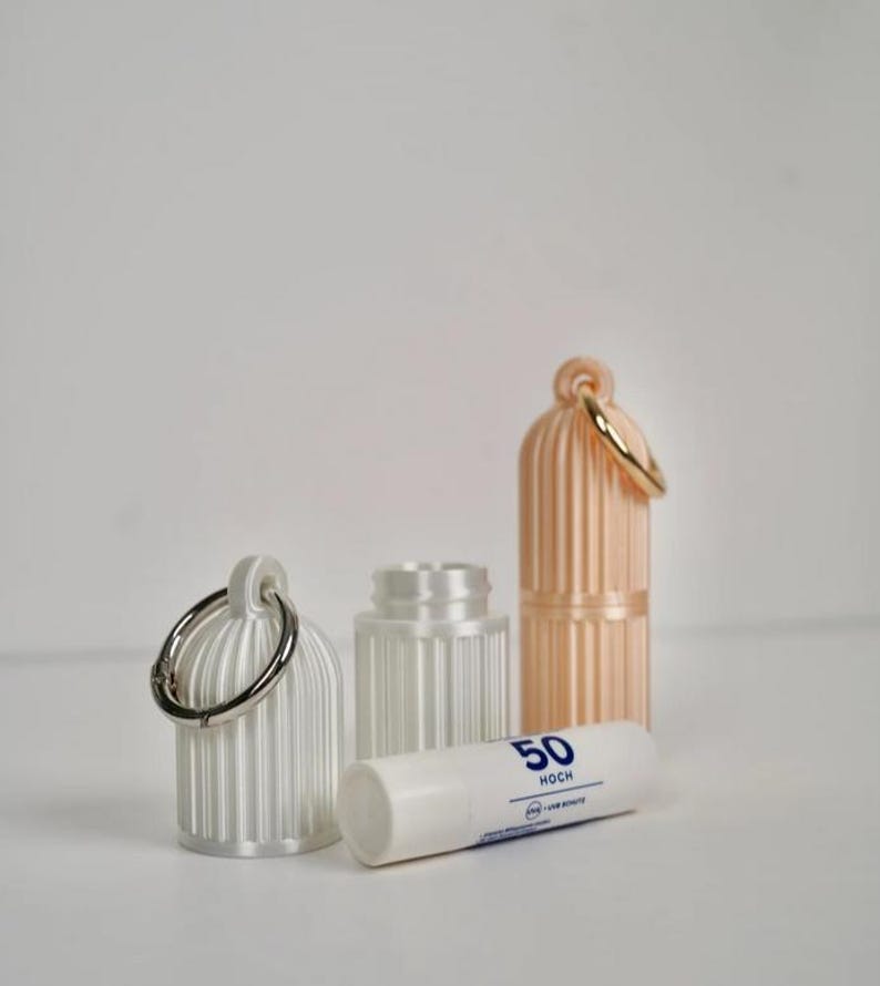 May include: Two cylindrical, ribbed, white and peach-colored plastic cases with attached metal rings, alongside a white lip balm tube labeled with the number 50 and the word HOCH, featuring a small blue icon and the text UVA + UVB SCHUTZ.