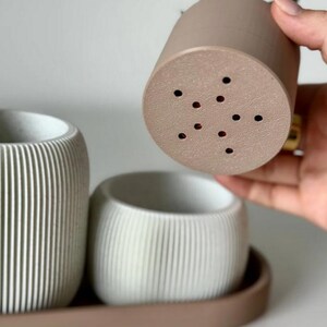 May include: A set of three modern, minimalist planters in shades of beige and white. The planters have a ribbed texture and are arranged on a matching tray. One planter is being held up, revealing drainage holes.