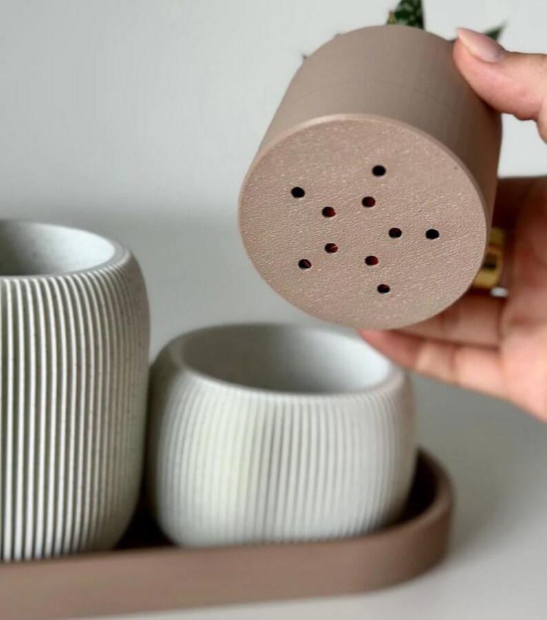 May include: A set of three modern, minimalist planters in shades of beige and white. The planters have a ribbed texture and are arranged on a matching tray. One planter is being held up, revealing drainage holes.