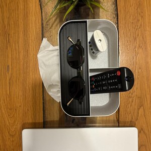 Op de afbeelding: Een houten tafel met een witte en zwarte organizer met zonnebril, afstandsbediening en een klein wit apparaat. Een zilveren laptop met een Apple-logo staat onderaan. Een kleine groene plant staat op de tafel.