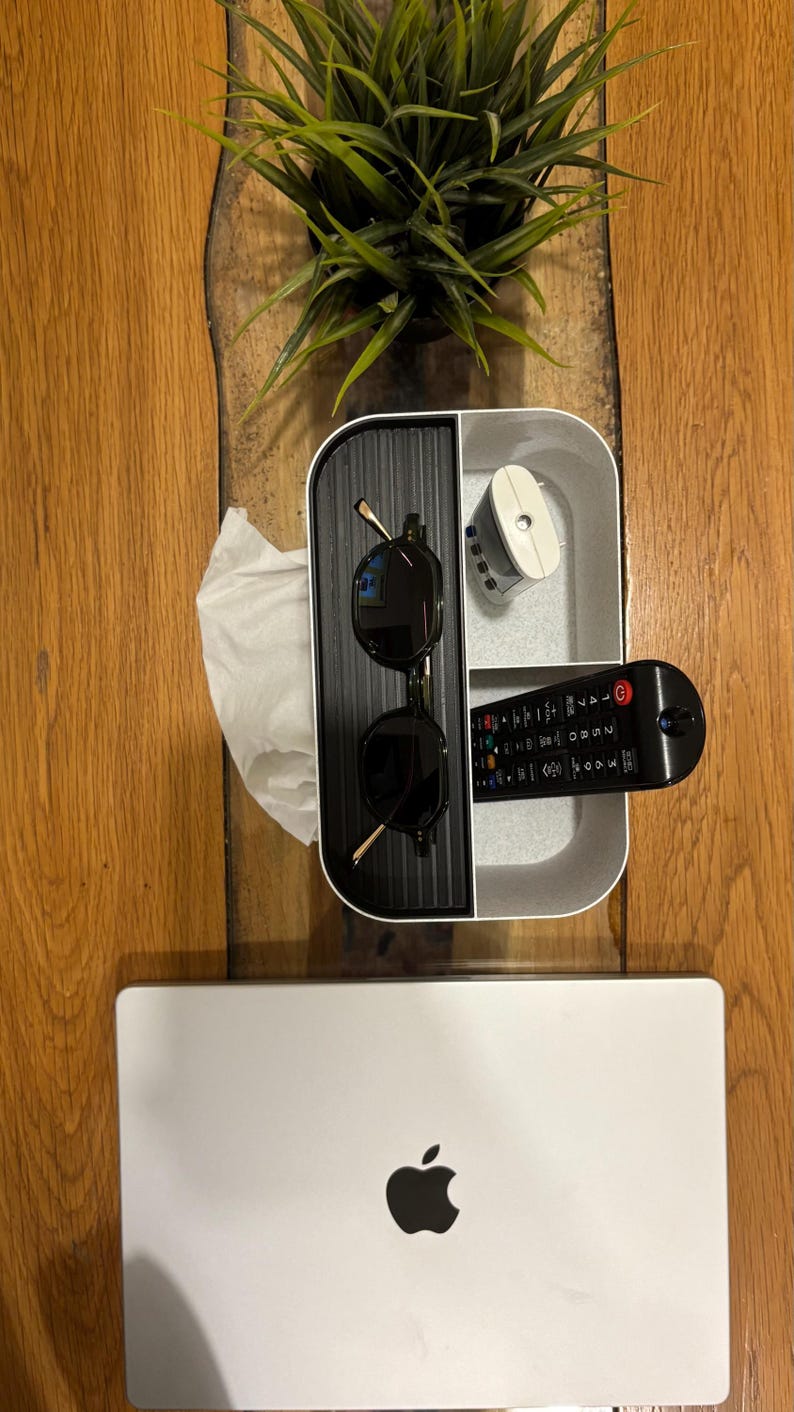 Op de afbeelding: Een houten tafel met een witte en zwarte organizer met zonnebril, afstandsbediening en een klein wit apparaat. Een zilveren laptop met een Apple-logo staat onderaan. Een kleine groene plant staat op de tafel.