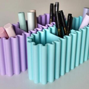 K&ouml;nnte beinhalten: Zwei pastellfarbene Schreibtisch-Organizer. Einer ist lavendelfarben, der andere hellblau. Jeder hat ein wellenf&ouml;rmiges Design und vertikale F&auml;cher. Die Organizer halten Stifte, Marker und andere Schreibwaren.