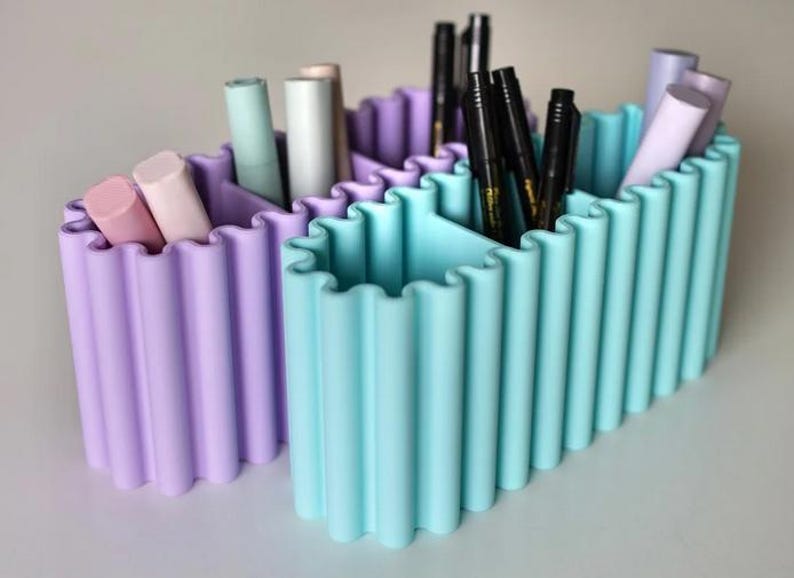 K&ouml;nnte beinhalten: Zwei pastellfarbene Schreibtisch-Organizer. Einer ist lavendelfarben, der andere hellblau. Jeder hat ein wellenf&ouml;rmiges Design und vertikale F&auml;cher. Die Organizer halten Stifte, Marker und andere Schreibwaren.