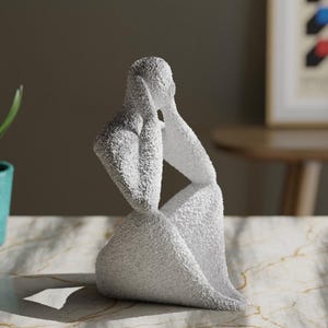 May include: A white, textured sculpture of a seated figure with hands raised to the sides of the head. The figure has a rough, granular surface finish and is depicted in a contemplative pose, with the body angled slightly to the side.