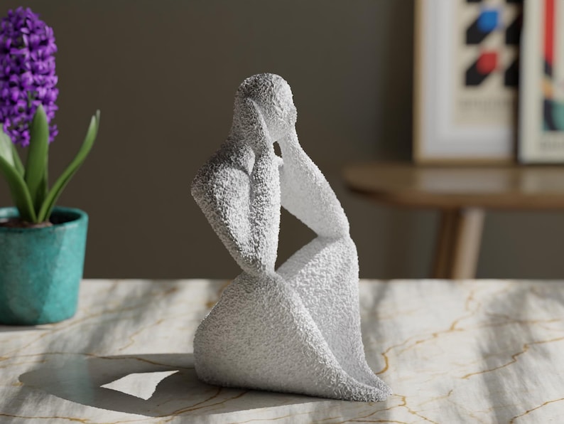 May include: A white, textured sculpture of a seated figure with hands raised to the sides of the head. The figure has a rough, granular surface finish and is depicted in a contemplative pose, with the body angled slightly to the side.