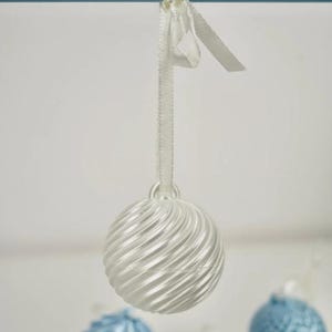 Pode incluir: Um ornamento esf&eacute;rico branco com uma textura canelada em espiral, suspenso por uma fita de cetim branca. O ornamento apresenta um acabamento brilhante e faz parte de uma cole&ccedil;&atilde;o de decora&ccedil;&otilde;es esf&eacute;ricas semelhantes em branco e azul claro.