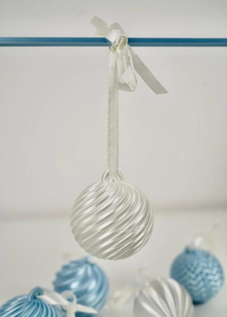 Pode incluir: Um ornamento esf&eacute;rico branco com uma textura canelada em espiral, suspenso por uma fita de cetim branca. O ornamento apresenta um acabamento brilhante e faz parte de uma cole&ccedil;&atilde;o de decora&ccedil;&otilde;es esf&eacute;ricas semelhantes em branco e azul claro.