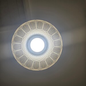 May include: A low-angle shot of a white, geometric-patterned light fixture with a bright, circular light source at its center. The fixture has a circular shape with a series of geometric shapes radiating outwards. The background is a neutral gray.