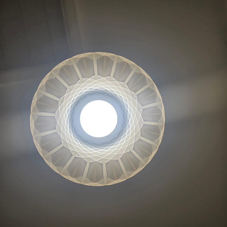 May include: A low-angle shot of a white, geometric-patterned light fixture with a bright, circular light source at its center. The fixture has a circular shape with a series of geometric shapes radiating outwards. The background is a neutral gray.
