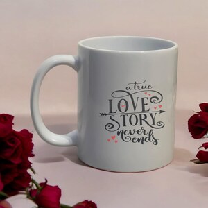 May include: White ceramic mug with a curved handle. The mug features the text "a true LOVE STORY never ends" in black script with pink heart accents. The mug is a standard size for coffee or tea.