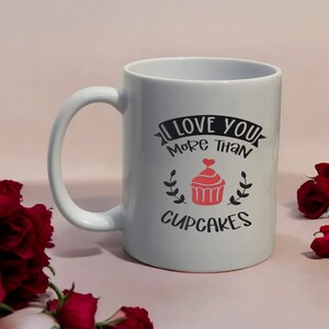 May include: White ceramic mug with a curved handle. The mug features the text "I LOVE YOU More Than CUPCAKES" in black, with a red cupcake graphic. The mug is surrounded by red roses.