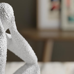 May include: A white sculpture of a seated figure with a textured, granular surface. The figure is depicted in a hunched, contemplative pose with arms raised toward the head, showcasing a rough, uneven finish across the entire form of the statue.