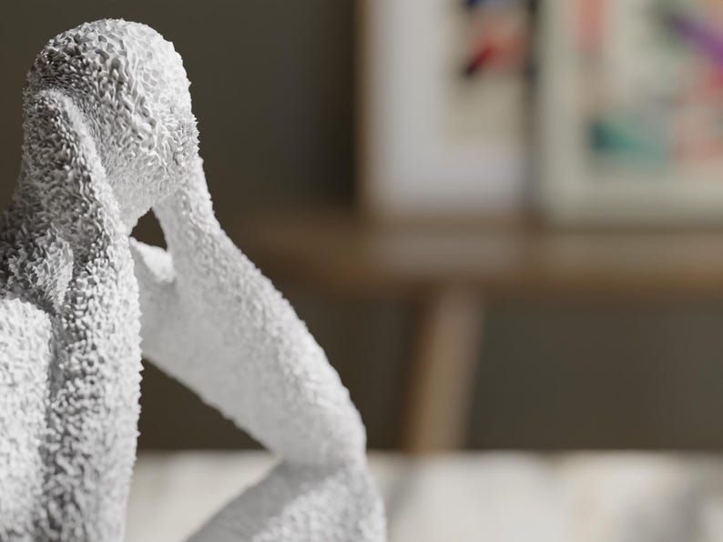 May include: A white sculpture of a seated figure with a textured, granular surface. The figure is depicted in a hunched, contemplative pose with arms raised toward the head, showcasing a rough, uneven finish across the entire form of the statue.