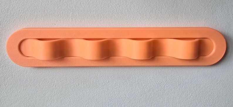 May include: A horizontal, rectangular, peach-colored wall-mounted organizer featuring four rounded, wave-like slots designed for holding items. The object has a smooth, matte finish and a uniform, solid color throughout its entire structure.