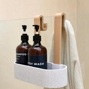 K&ouml;nnte beinhalten: Ein hellgrauer, gerippter, ovaler Duschkorb mit zwei beigefarbenen Haken zur Befestigung an einer Glaswand. Der Korb enth&auml;lt zwei dunkelbraune Flaschen mit schwarzen Pumpspendern, beschriftet mit Shampoo und Body Wash, sowie ein beiges Handtuch.
