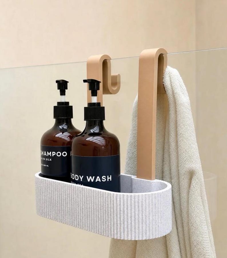 K&ouml;nnte beinhalten: Ein hellgrauer, gerippter, ovaler Duschkorb mit zwei beigefarbenen Haken zur Befestigung an einer Glaswand. Der Korb enth&auml;lt zwei dunkelbraune Flaschen mit schwarzen Pumpspendern, beschriftet mit Shampoo und Body Wash, sowie ein beiges Handtuch.
