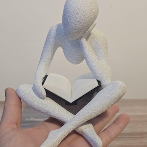 May include: A white, textured sculpture of a person reading a book while sitting cross-legged. The figure is holding the book open with both hands, with the head resting on one hand. The sculpture has a minimalist design.