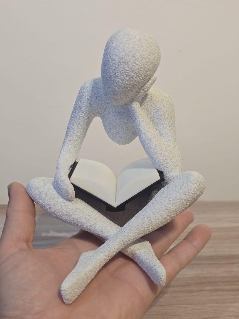 May include: A white, textured sculpture of a person reading a book while sitting cross-legged. The figure is holding the book open with both hands, with the head resting on one hand. The sculpture has a minimalist design.