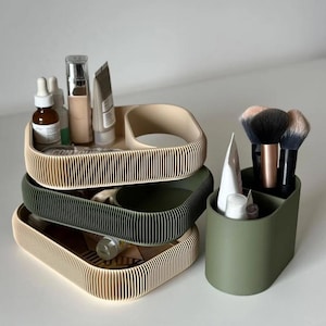 Op de afbeelding: Een stapel van drie beige en groene geribbelde cosmeticalades met diverse schoonheidsproducten. Een aparte olijfgroene container bevat make-up borstels en tubes. De trays zijn rechthoekig met afgeronde hoeken.