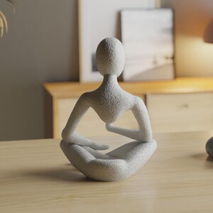 Op de afbeelding: Een wit, getextureerd beeldhouwwerk van een gestileerde menselijke figuur in een zittende, meditatieve houding. De figuur heeft een glad, ovaalvormig hoofd, brede schouders en armen die op gekruiste benen rusten. Het oppervlak is korrelig.