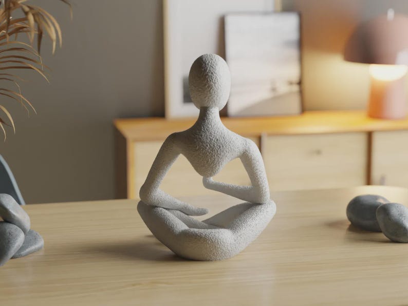 Op de afbeelding: Een wit, getextureerd beeldhouwwerk van een gestileerde menselijke figuur in een zittende, meditatieve houding. De figuur heeft een glad, ovaalvormig hoofd, brede schouders en armen die op gekruiste benen rusten. Het oppervlak is korrelig.