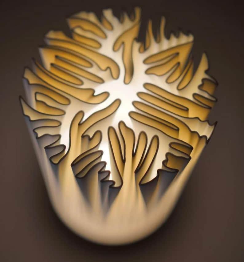 Puede incluir: Una l&aacute;mpara abstracta con un dise&ntilde;o en capas. Presenta formas org&aacute;nicas curvas en tonos crema y dorado, delineadas en negro. La fuente de luz interna crea un brillo suave. La forma general recuerda a una flor.
