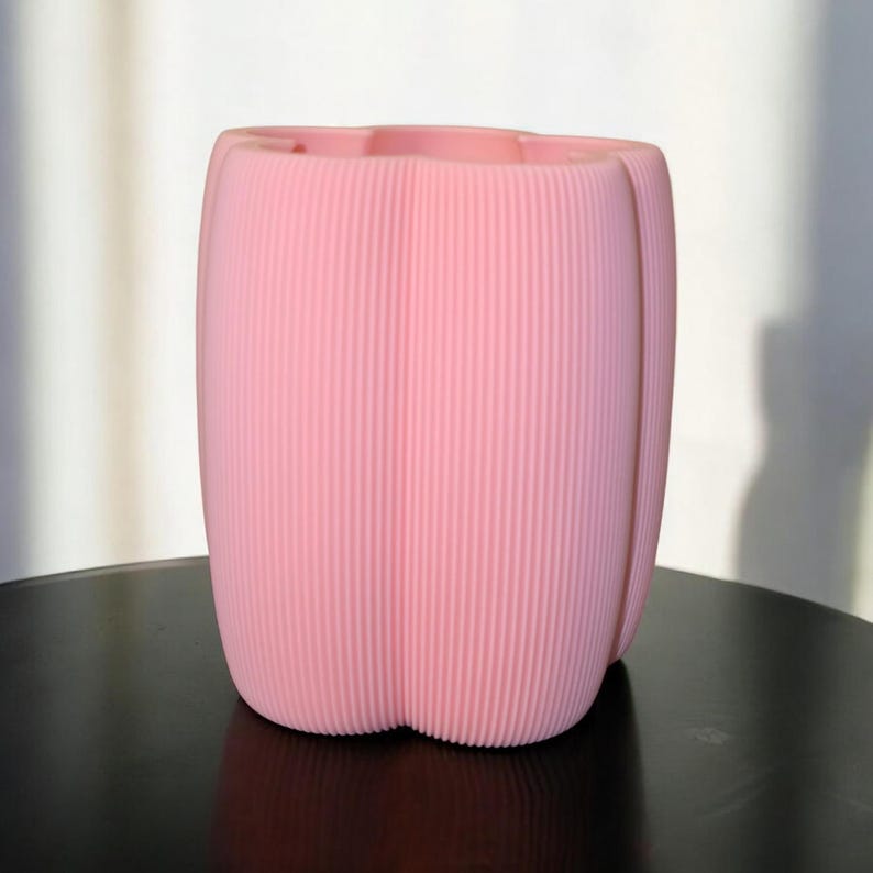 May include: A pink, ribbed planter with a tulip-like shape. The vertical ridges create a textured surface. The planter sits on a dark surface, and is illuminated by natural light.